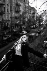 Black and white photo of stylish woman posing on the balcony in the city. Spring autumn fashion concept. Fashionable woman resting on terrace.