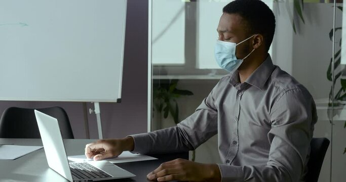 Upset young African business man sits in front of laptop, wears medical mask, reads online bad news about continuation of quarantine outbreak of covid virus, feels headache and stress, concept of loss