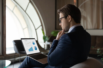 Side back view focused skilled young 30s male marketing specialist in suit analyzing graphs and...