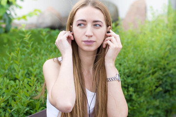 Girl with blond hair sitting outdoors in beautiful green park listening music with headphones, close-up portrait