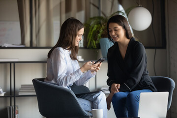 Happy friendly mixed race female colleagues enjoying break pause time in office, watching funny video on phone, enjoying informal conversation communication together indoors, work relations concept.