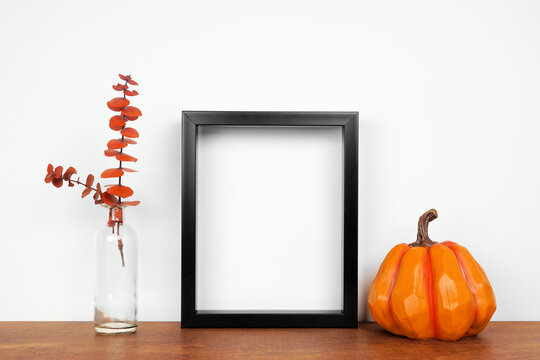 Mock up black frame with fall branches and pumpkin decor on a wood shelf. Autumn concept. Portrait frame against a white wall.