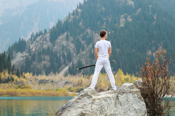 A young man stands on a large stone and holds a Japanese sword in his hands.