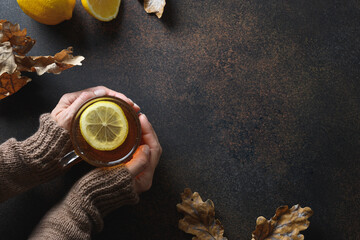 Warming tea with lemon in womans hands in knitting sweater on brown background. View from above with copy space.