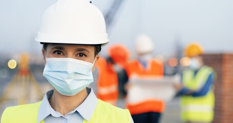 Fototapeta na wymiar Portrait of female builder or architect wearing protective helmet in construction site. Woman engineer in medical face mask looking at camera.