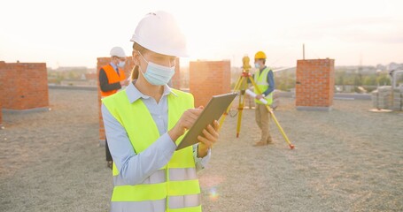 Caucasian young good-looking woman engineer in helmet and medical mask standing at constructing side, using tablet device and tapping on screen. Outdoors. Female constructor with gadget computer.