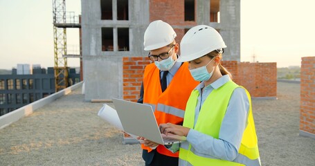 Caucasian couple of male and female constructors in helmets and medical masks talking and discussing plan draft at building side. Man and woman builders working at constructing with laptop computer.