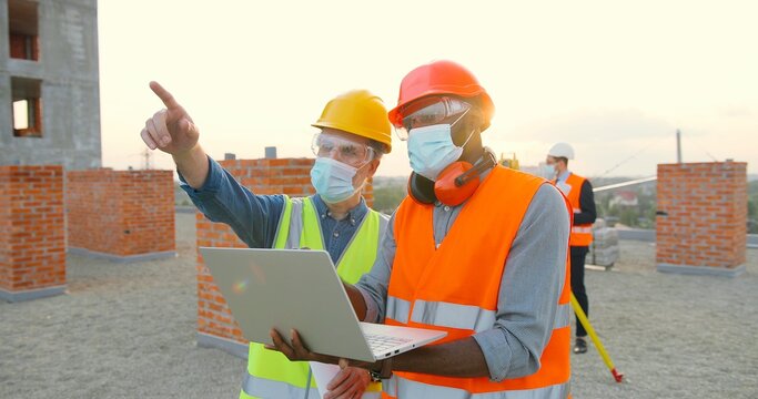 Group Of Builders In Hardhat Works On The Building Site. Two Male Builders In Medical Face Mask Using Laptop Computer On Construction Site.