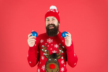 New year concept. Christmas party. Winter outfit. Good vibes. Winter decorations. Fun festive activities. Hipster smiling cheerful bearded man wear winter sweater and hat hold balls red background