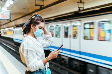 Woman with protective mask on face using smart phone while standing at subway station