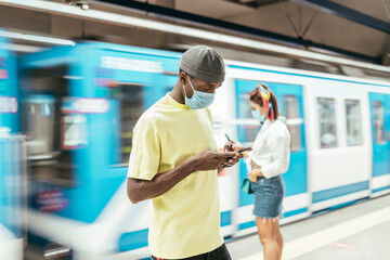 Fototapeta na wymiar Black man with protective mask on face using smart phone while standing against subway in motion