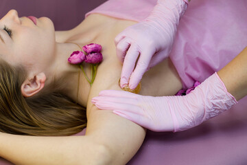 a young woman is shugaring her armpits. purple background. The master applies a paste for sugaring