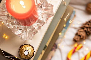 
Christmas or New Year's atmosphere. Composition with a stack of books, a red candle, seasonal sweets and garland lights. Home atmosphere, relaxation, winter holidays