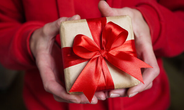 Young woman in red swether holding a gift box wrapped in brown paper and tied with a red ribbon with a bow for a space event. Christmas, New Year, Birthday, Valentine's Day.Close-up