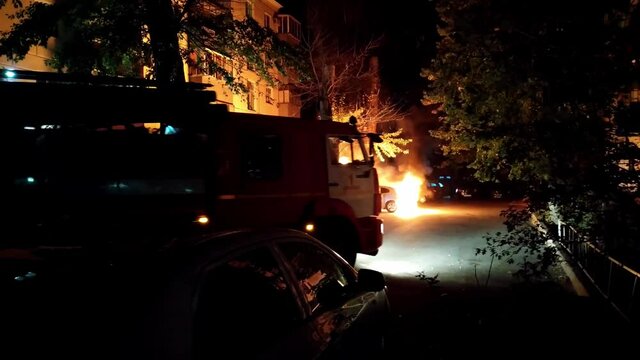 Fire truck arrives to extinguish the arson of car terrorism in a residential area. Theme of vandalism and hooliganism at night. Automobile engulfed in flames of fire. Trees close to burning sedan.
