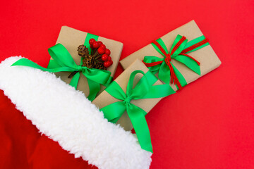 Gifts wrapped in craft paper and decorated with green and red ribbons flat lay on red background next to a Christmas hat
