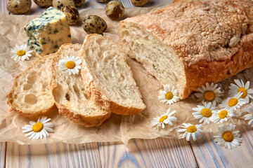 Homemade ciabatta in baking paper, blue gorgonzola cheese, olive and quail eggs. Chamomile flowers. Wooden rustic background.