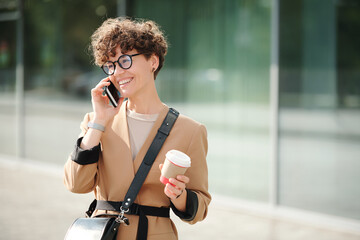 Fototapeta premium Young cheerful businesswoman with drink in hand and smartphone by ear