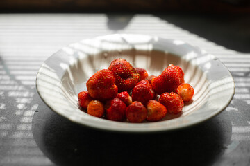 picture of juicy fresh ripe red strawberries in a white ceramic plate on the table under bright sunlight in a village