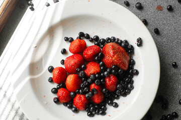 Top view stock photo of sweet and tasty juicy strawberries and black currant in ceramic plate. Some berries are scattered on the table.