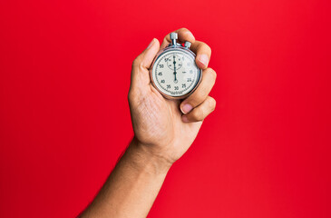 Hand of young hispanic man using stopwatch over isolated red background.