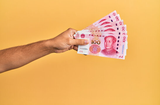 Hand of hispanic man holding chinese 100 yuan banknotes over isolated yellow background.