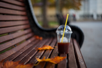 A clear glass of coffee with a yellow tube stands on a wooden bench. Soft and selective focus. Autumn leaves in the background.