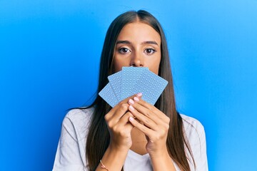 Young hispanic woman playing gambling poker covering face with cards relaxed with serious expression on face. simple and natural looking at the camera.
