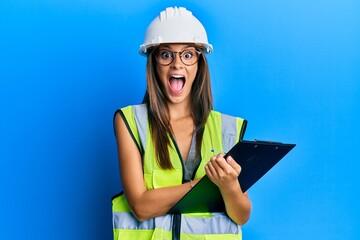 Young hispanic woman wearing safety helmet holding clipboard celebrating crazy and amazed for success with open eyes screaming excited.