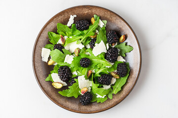 Fresh salad with arugula, goat cheese, blackberries and pistachios in a plate on white background. top view