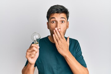 Young handsome man holding lightbulb for inspiration and idea covering mouth with hand, shocked and afraid for mistake. surprised expression