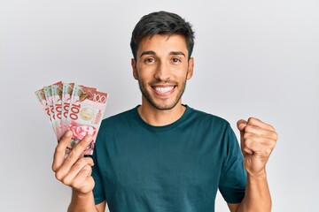 Young handsome man holding 100 new zealand dollars banknote screaming proud, celebrating victory and success very excited with raised arm