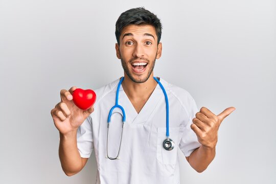 Young handsome man wearing doctor uniform holding heart pointing thumb up to the side smiling happy with open mouth