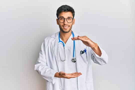 Young handsome man wearing doctor uniform and stethoscope gesturing with hands showing big and large size sign, measure symbol. smiling looking at the camera. measuring concept.