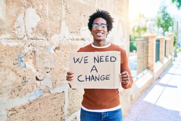 Naklejka na ściany i meble Young handsome african american man asking for change smiling happy. Standing with smile on face holding we need a change banner cardboard at town street.