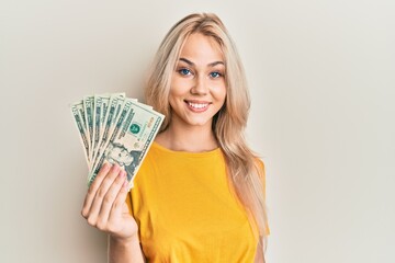 Beautiful caucasian blonde girl holding 20 dollars banknotes looking positive and happy standing and smiling with a confident smile showing teeth