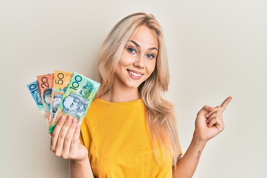 Beautiful caucasian blonde girl holding australian dollars smiling happy pointing with hand and finger to the side