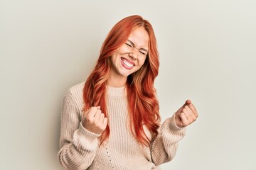 Young beautiful redhead woman wearing casual winter sweater very happy and excited doing winner gesture with arms raised, smiling and screaming for success. celebration concept.