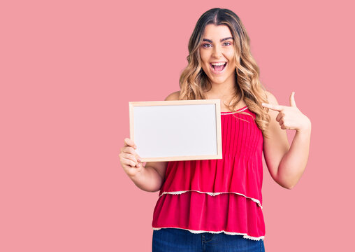 Young caucasian woman holding empty white chalkboard pointing finger to one self smiling happy and proud