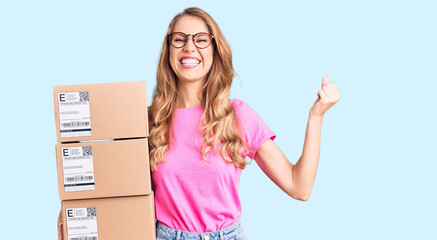 Young caucasian woman with blond hair holding delivery package screaming proud, celebrating victory and success very excited with raised arms