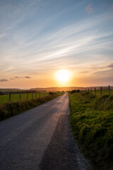 a single lane road in the british country side leading into a sunset