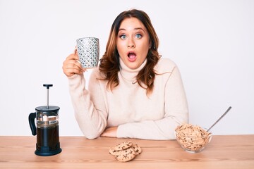 Young beautiful caucasian woman sitting on the table having breakfast scared and amazed with open...