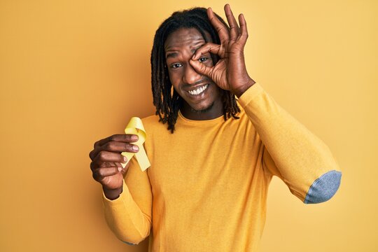 African american man with braids holding suicide prevention yellow ribbon smiling happy doing ok sign with hand on eye looking through fingers