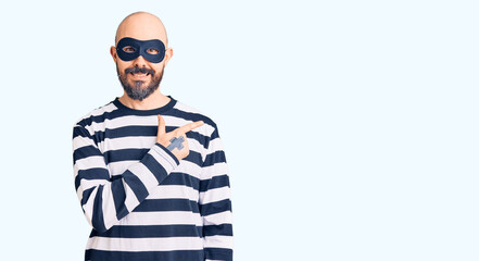 Young handsome man wearing burglar mask cheerful with a smile on face pointing with hand and finger up to the side with happy and natural expression