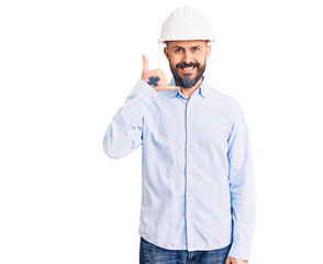Young handsome man wearing architect hardhat smiling doing phone gesture with hand and fingers like talking on the telephone. communicating concepts.