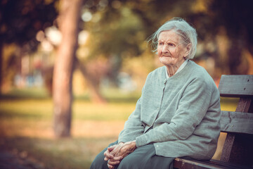 Portrait of senior woman sitting on bench in autumn park. Old lady feeling lonely and sad. - obrazy, fototapety, plakaty