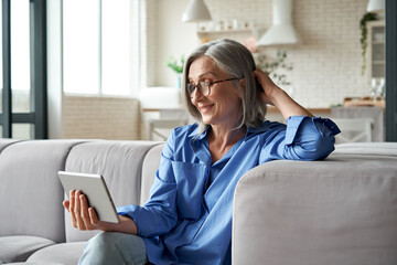 Happy 60s older mature middle aged adult woman holding digital tablet computer conference calling by social distance virtual family online chat meeting or watching video sitting on couch at home.