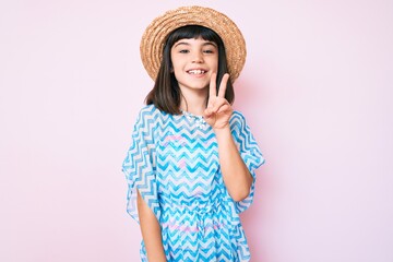 Young little girl with bang wearing summer dress and hat smiling with happy face winking at the camera doing victory sign. number two.