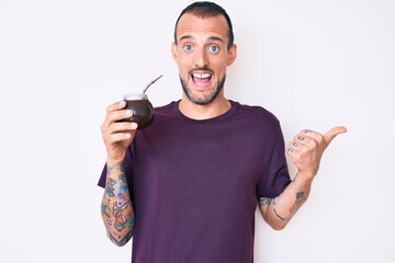 Young handsome man with tattoo drinking mate infusion pointing thumb up to the side smiling happy...