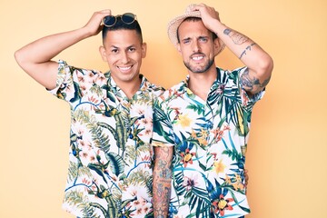 Young gay couple of two men wearing summer hat and hawaiian shirt smiling confident touching hair with hand up gesture, posing attractive and fashionable